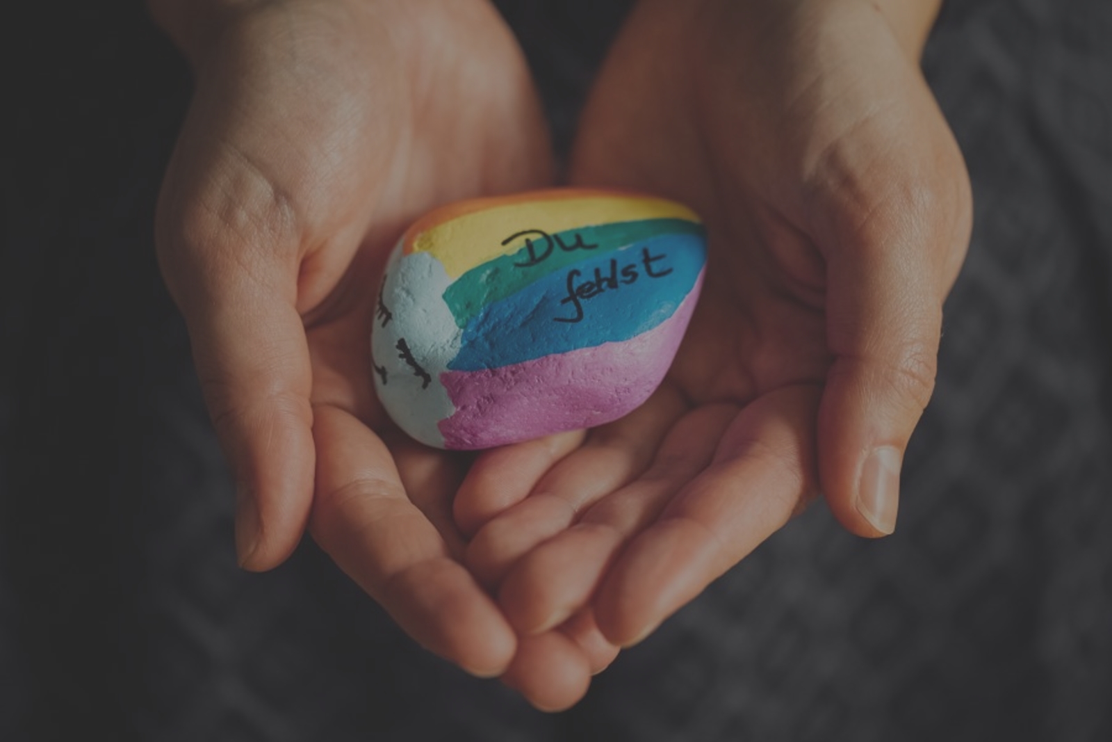 Zwei Hände halten einen Regenbogen-Stein mit der Aufschrift "Du fehlst"